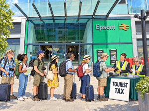 Tourists queuing outside Epsom Railway Station to pay their tax. Cartoon.